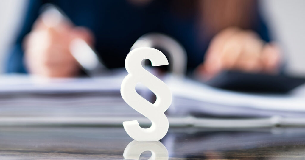 Foto: Paragraphenzeichen, dahinter unscharf Mann am Schreibtisch